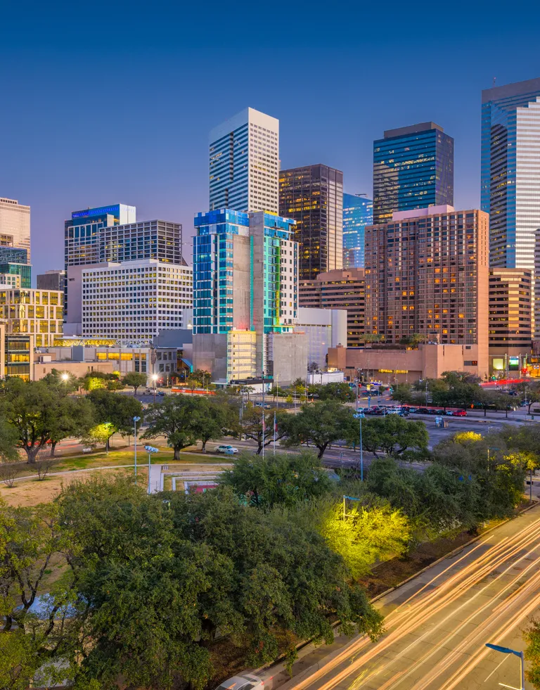 Evening cityscape of a downtown area with illuminated modern skyscrapers and a tree-lined street with light trails.