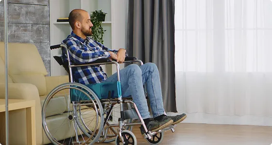 Man in blue plaid shirt sitting in a wheelchair near window with curtains.