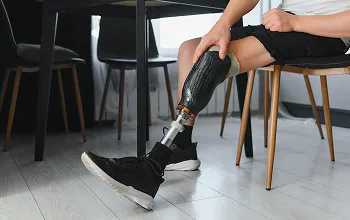 Person adjusting a black prosthetic leg while seated indoors on a wooden chair.