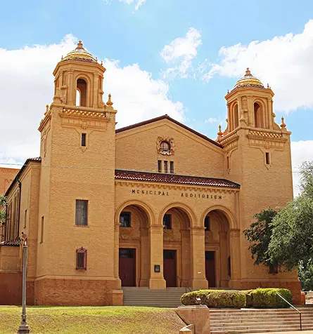 Exterior view of a tan brick municipal auditorium with two symmetrical towers under a partly cloudy sky.