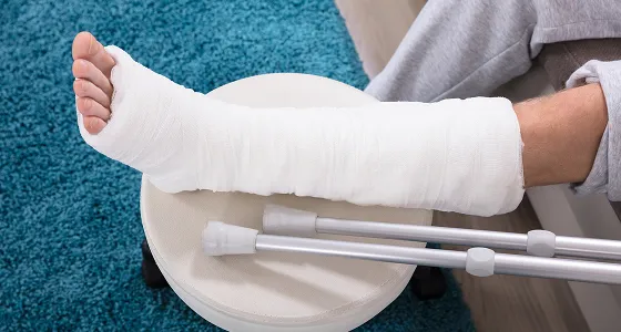 Person's leg in a white cast resting on a stool with crutches nearby.