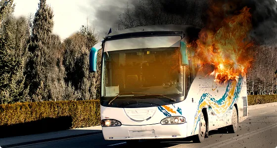 White and blue bus on fire with flames and smoke on right side, roadside trees visible.