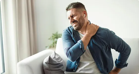 Middle-aged man in blue shirt sitting on white couch, holding neck in pain.