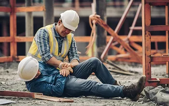 Two construction workers in safety helmets and vests, one assisting the other on the ground.