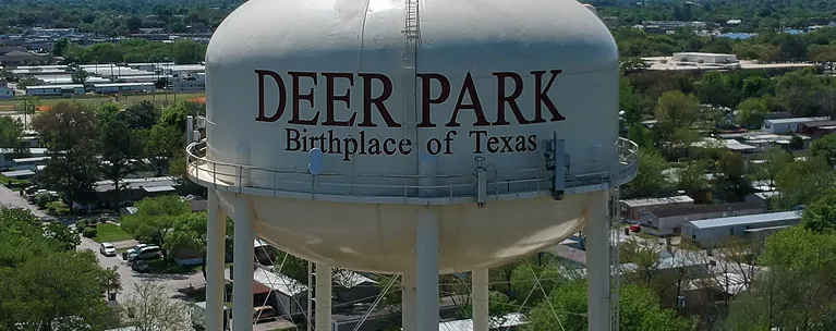 Large white water tower with "DEER PARK Birthplace of Texas" text, surrounded by trees and buildings.
