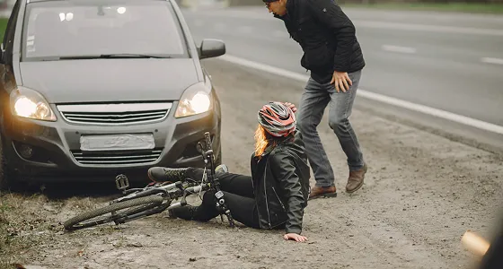 Woman wearing a helmet sitting on the ground next to a fallen bicycle and a car, with a man standing nearby.