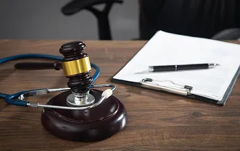 A wooden gavel, stethoscope, and clipboard with pen on a wooden desk.
