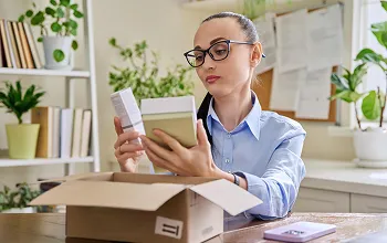 Woman in glasses unpacking and examining products from a cardboard box at a desk.
