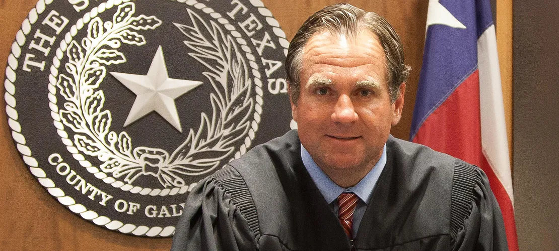 Middle-aged male judge in black robe sitting before Texas county seal and flag.