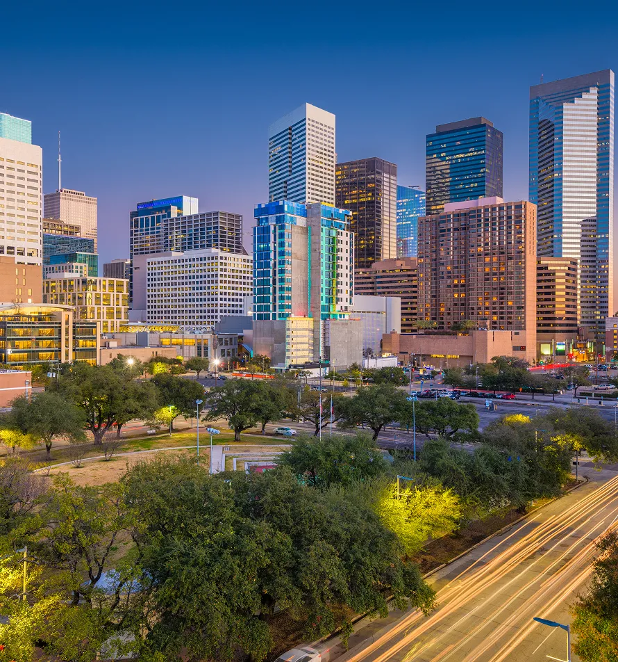 Dusk view of a city skyline with illuminated skyscrapers, green trees, and light trails on a busy road.