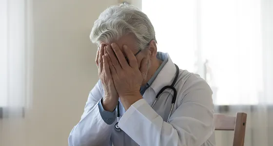 Older male doctor in white coat with stethoscope covering his face with hands.