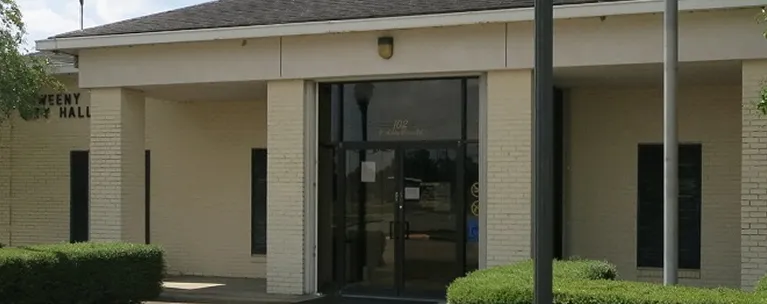Entrance of a beige brick building with glass double doors and surrounding greenery.