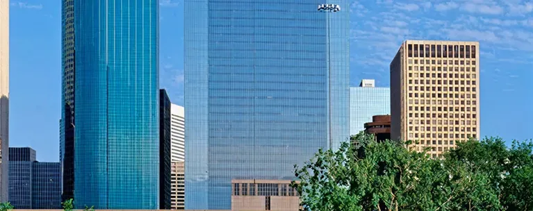 Panoramic view of modern glass and concrete skyscrapers with green trees in foreground.
