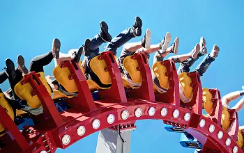People with raised legs riding a red and yellow roller coaster against a clear blue sky.