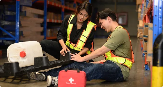 Two warehouse workers wearing yellow safety vests attending to a leg injury, with a red first aid kit nearby.