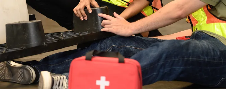 Two individuals in safety vests attending to a person lying on the ground with a red first aid kit nearby.