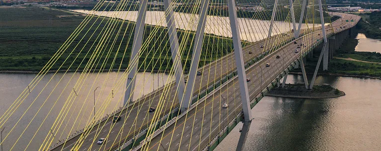 Aerial view of a modern cable-stayed bridge with yellow cables spanning over a river with vehicles on the roadway.