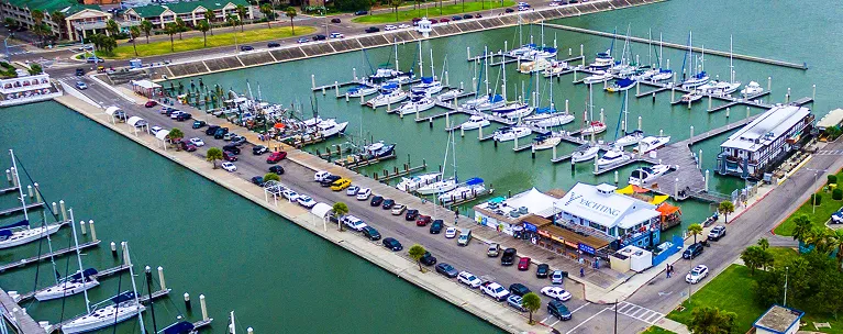 Aerial view of a marina with numerous docked sailboats and yachts, adjacent parking lot, and surrounding green areas.