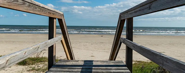 Wooden boardwalk railing leading to sandy beach with ocean waves under blue sky.