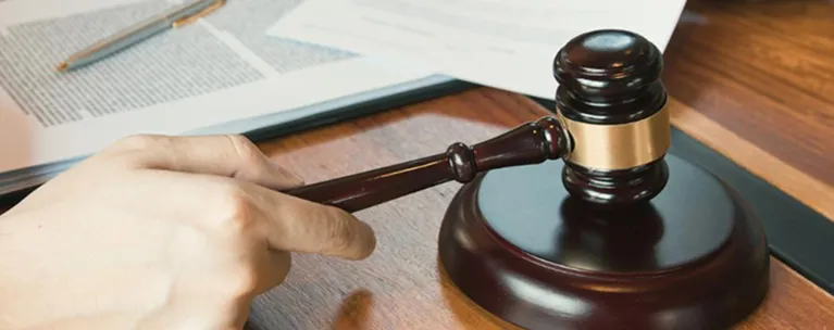 Hand holding a dark wooden gavel with golden band, documents and pen on table.