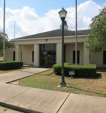 Exterior view of a beige brick town hall building with a central entrance and surrounding greenery.