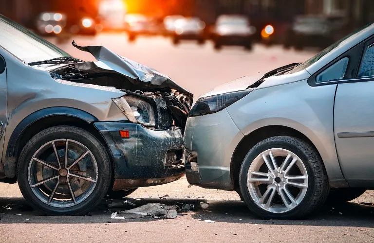 Two silver cars involved in a front-end collision on a city street during sunset.