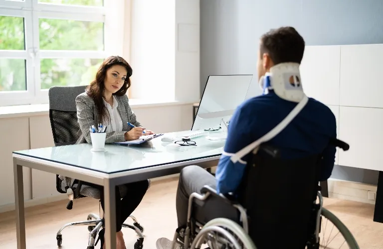 A professional woman in an office consulting a man in a wheelchair wearing a neck brace.