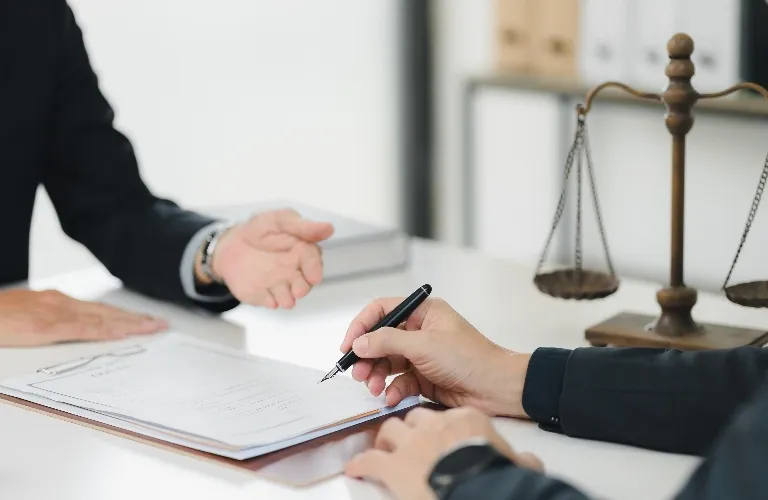 Two individuals in formal attire engaged in signing a document, with a wooden balance scale on the desk.