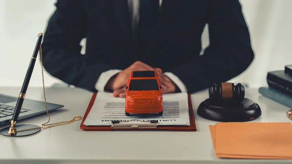 Person in suit behind desk with toy car, contract, gavel, and pen on white surface.