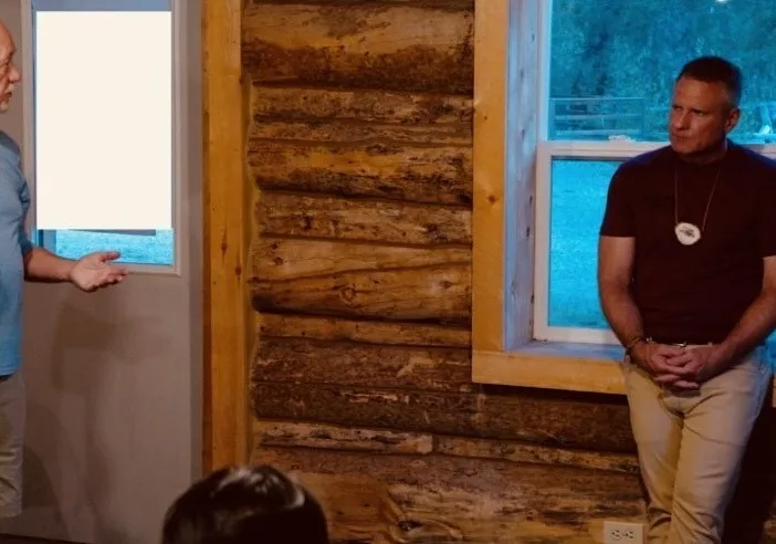 Two men engaged in conversation inside a rustic wooden cabin with large windows.