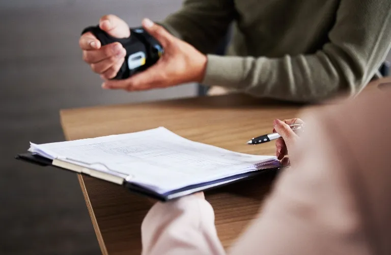Two people at a table, one holding a pen and clipboard, the other adjusting a wrist blood pressure monitor.