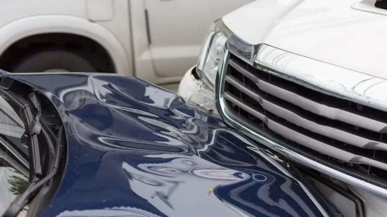 Close-up of a blue car with a severely dented hood after colliding with the front grille of a white vehicle.