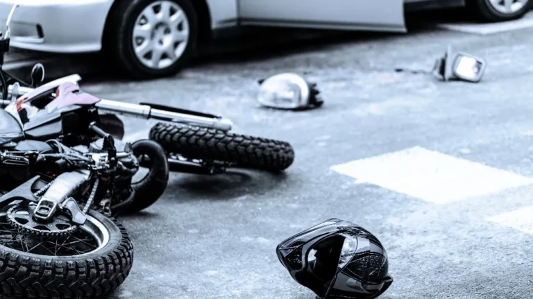 Black motorcycle lying on road after accident with detached helmet and car in background.