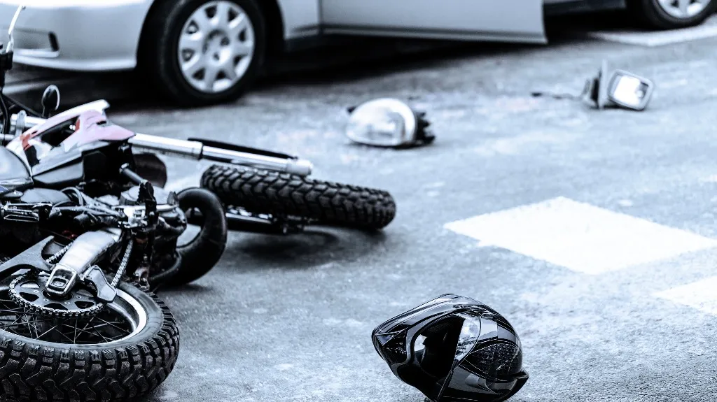 Black motorcycle lying on road after accident with detached helmet and car in background.
