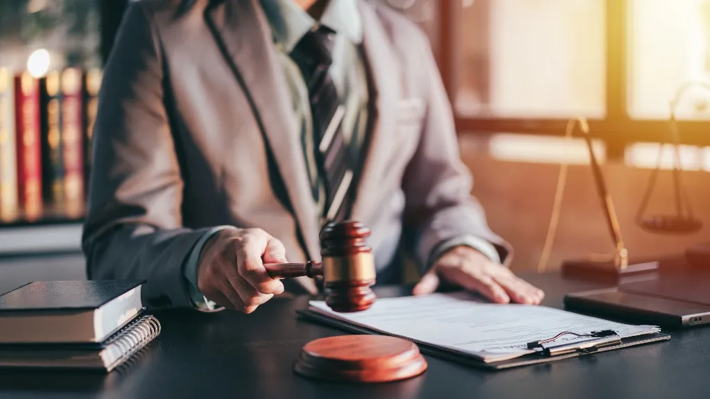 Person in a suit holding a wooden gavel over documents on a desk with legal books.