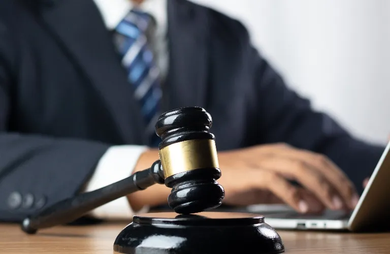 Close-up of a black judge’s gavel with a gold band, blurred person in suit typing on laptop.