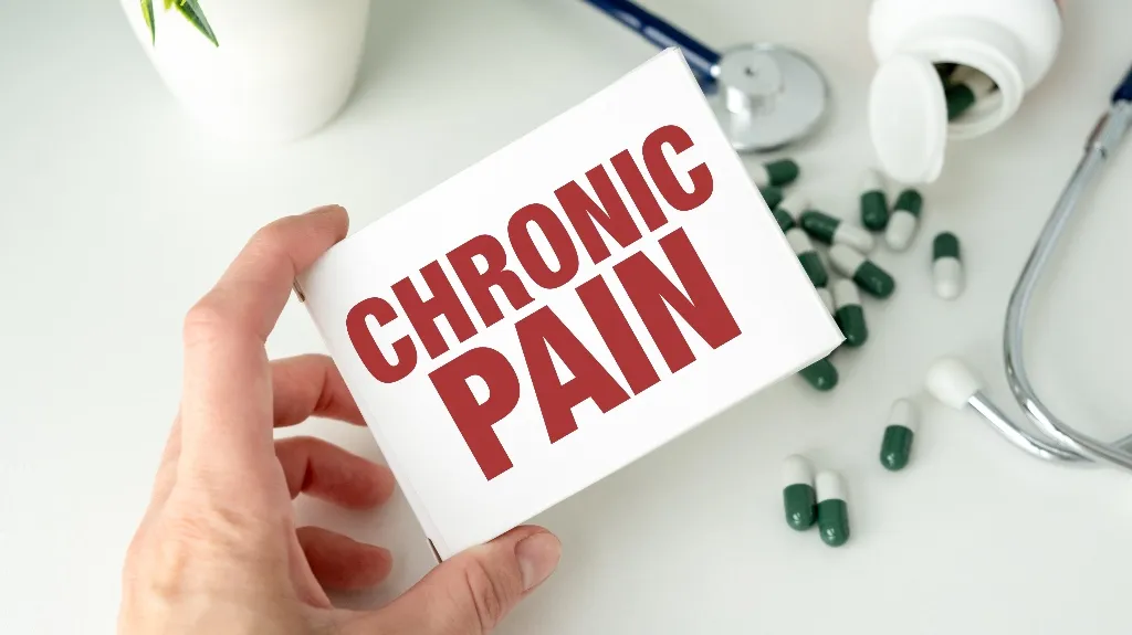 Hand holding a white box labeled "CHRONIC PAIN" in red, with green and white capsules, a stethoscope, and a white pill bottle on a white surface.