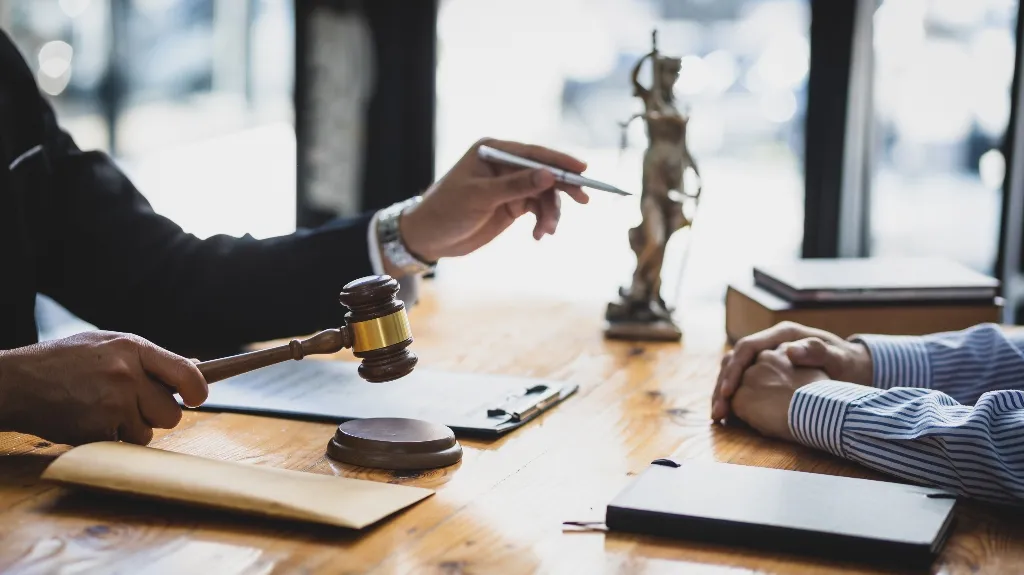 Two people at a wooden desk with a judge's gavel, legal books, and a Lady Justice statue.