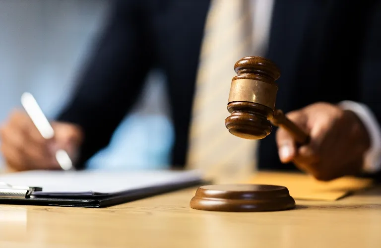 Close-up of a wooden judge's gavel being struck on a sound block, with a person writing in the background.