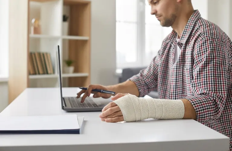 Man with a bandaged wrist working on a laptop at a white desk in an office.