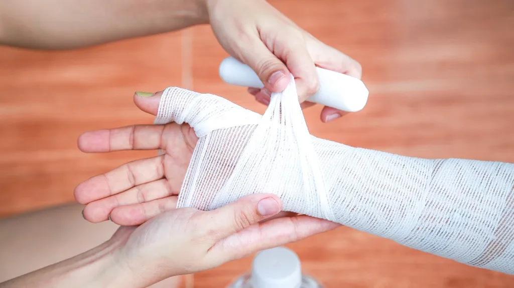 Person's hand being wrapped with white medical gauze bandage against a wooden floor background.