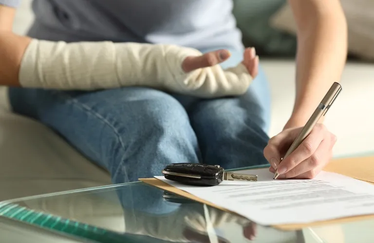 Person with a bandaged arm signing a document on a glass table with car keys nearby.
