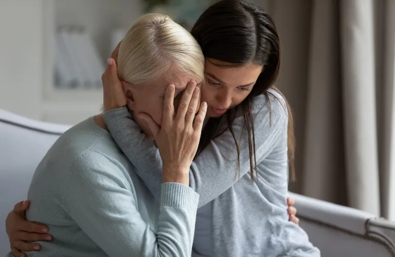 Two women in light gray sweaters, one comforting the other who is distressed and covering her face.
