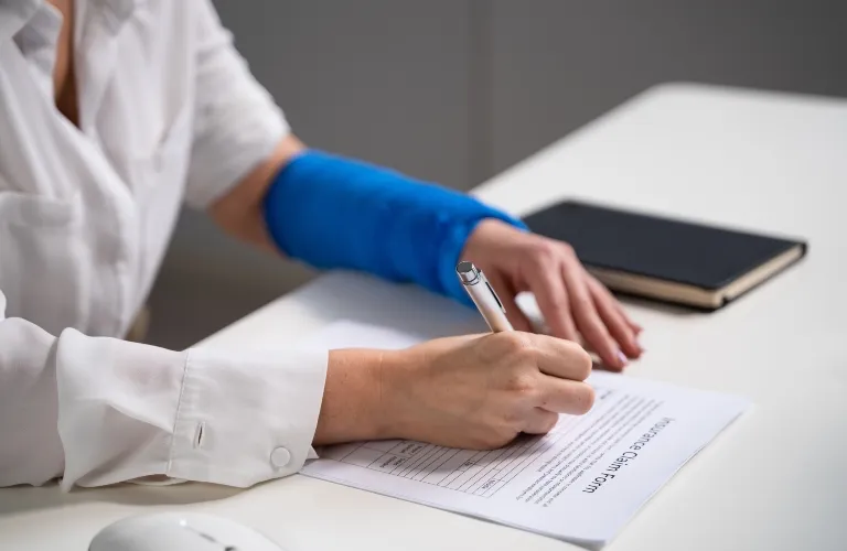 Person with blue arm cast filling out an insurance claim form on white desk.