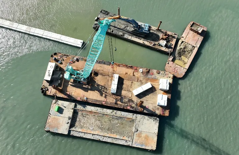 Aerial view of a floating construction barge with a blue crane and concrete blocks on murky water.