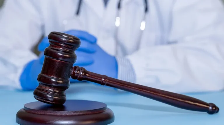 Wooden judge’s gavel in foreground with person wearing blue gloves and white coat in background.