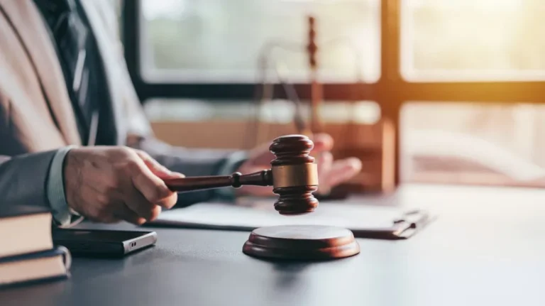 Person in suit holding wooden judge gavel above sounding block on desk.