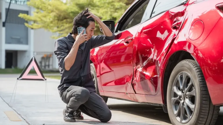 Man in dark clothing kneeling beside a red car with a large dent, making a phone call.
