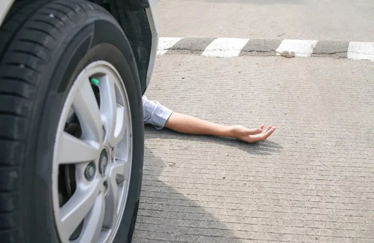 Close-up of a car tire on a road with a person's arm extended on the pavement nearby.