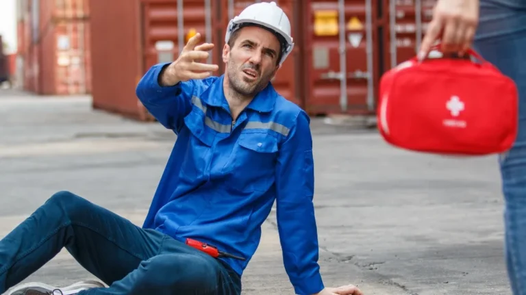 Man in blue work uniform and white hard hat sitting on ground, reaching out, red first aid kit visible.